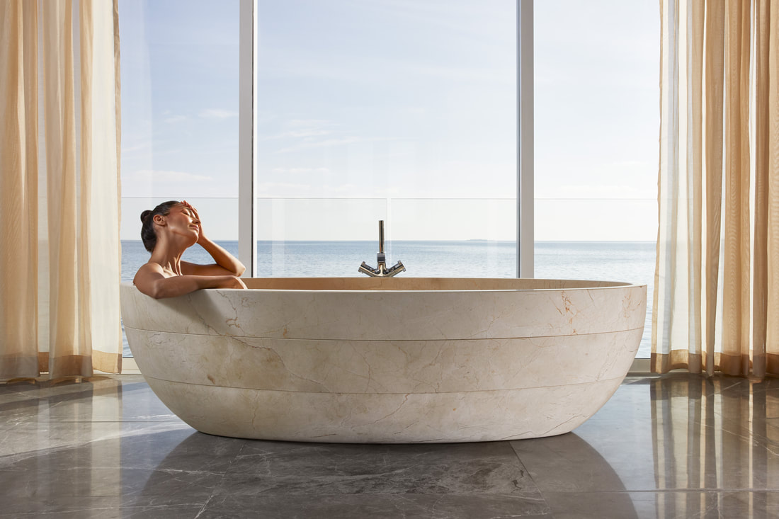 A tiered earthy marble soaking tub in the suites modern bathroom. Through the windows beyond the spa lays the uninterrupted ocean and clear blue sky.