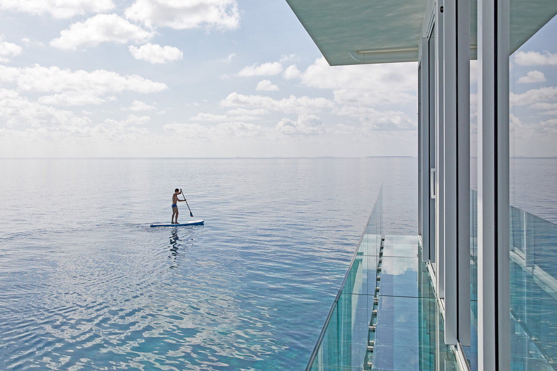 Right of frame is a the glass balcony of the over water villa with views of the ocean below and beyond. To the left of frame is a villa guest paddle boarding on the open Maldivian ocean.