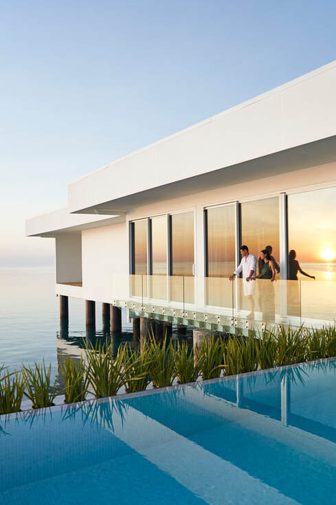 An infinity pool in the foreground with the white over water luxury villa in the background. The sunset can be seen through the reflection of the villas floor to ceiling windows.