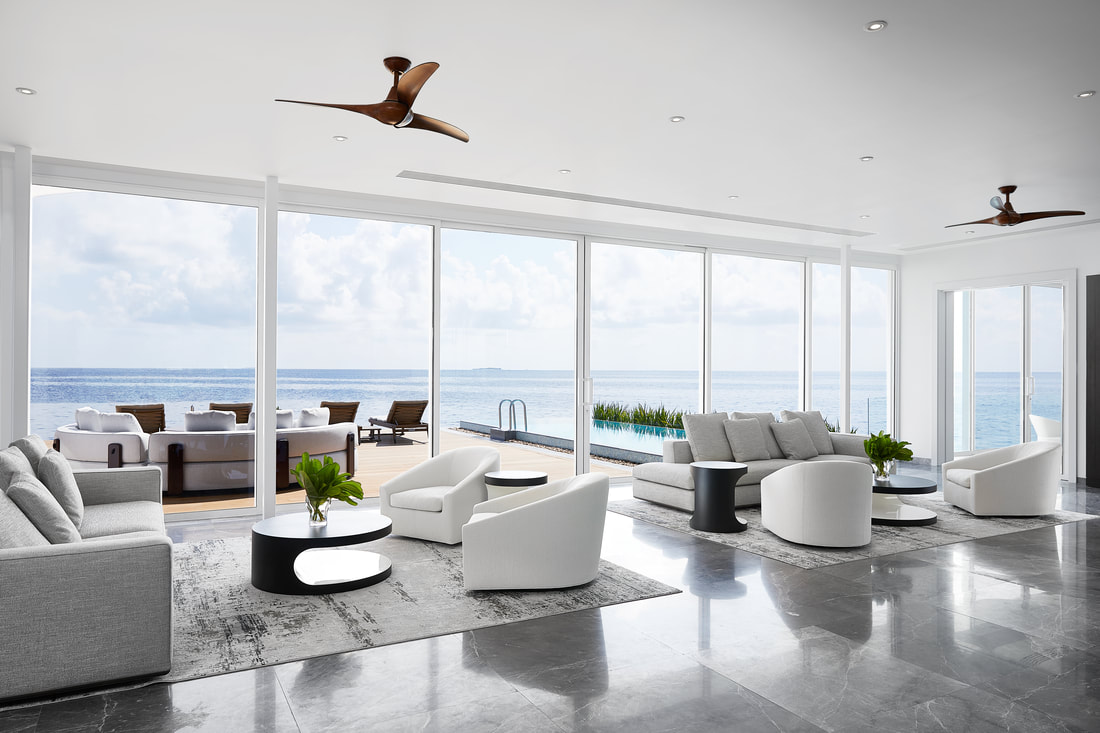 Modern living room with white marble bar of the over water villa. The gathering space has unobstructed views of the open Maldivian ocean. Beyond the sliding glass doors on the warm wood deck is an infinity pool and sun loungers.